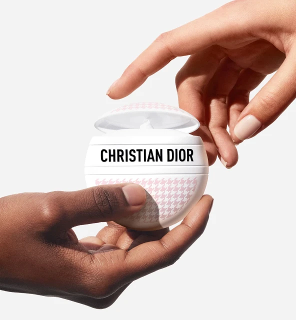 Christian Dior cream jar with a pink houndstooth pattern held by two hands against a white backdrop, close-up product shot.