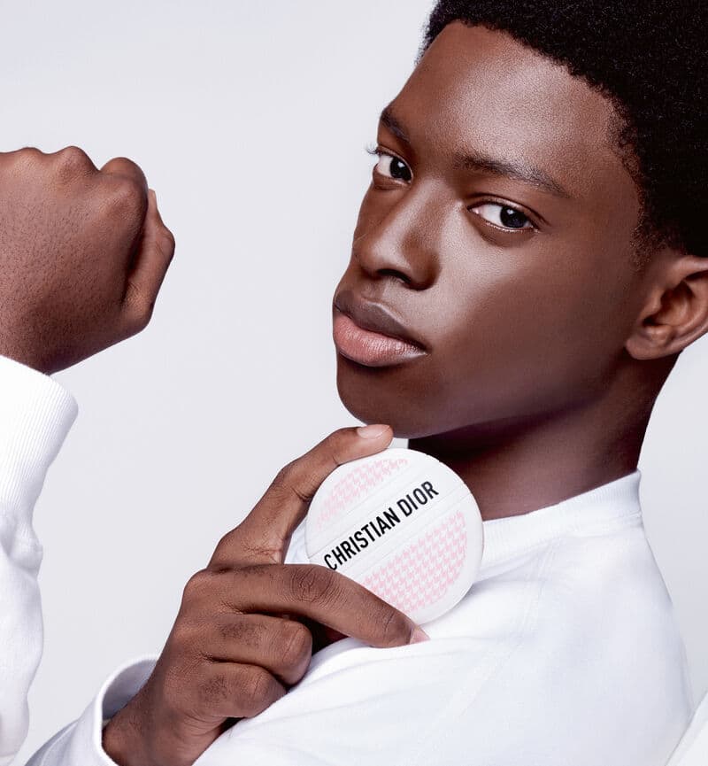 Close-up of a young adult Black male model holding a Christian Dior makeup compact, showcasing beauty and fashion in a clean photo studio setting.
