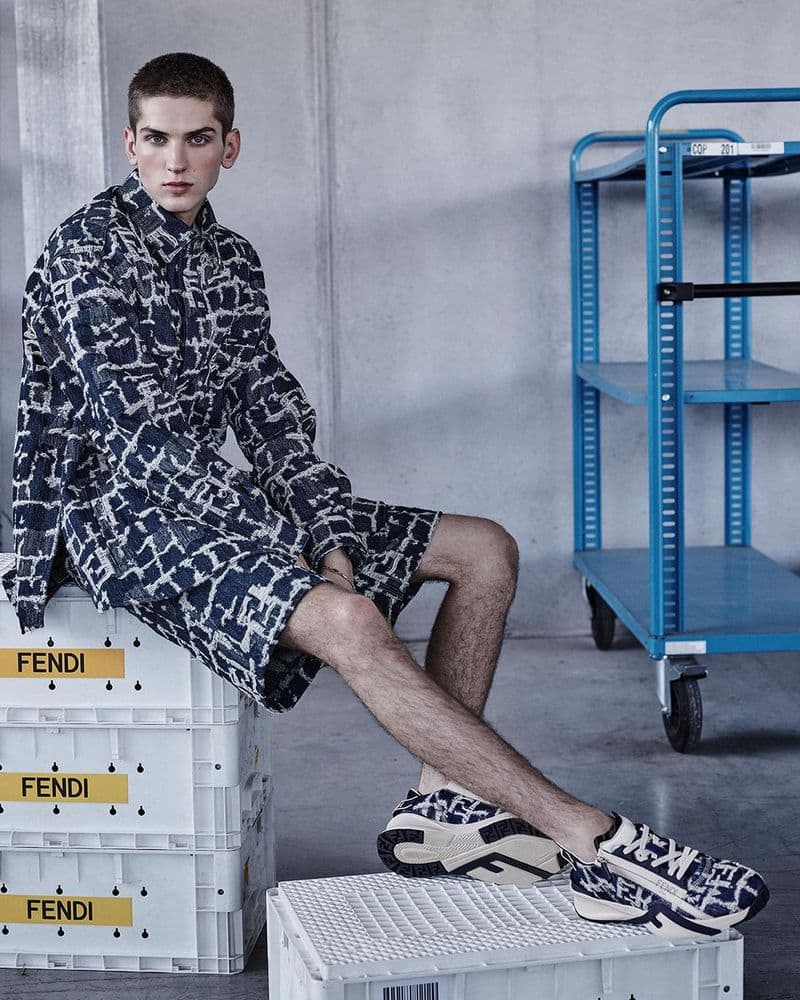 A young man poses in designer shirt and shorts sitting on branded white boxes in a studio, showcasing sneakers and a blue trolley in background.