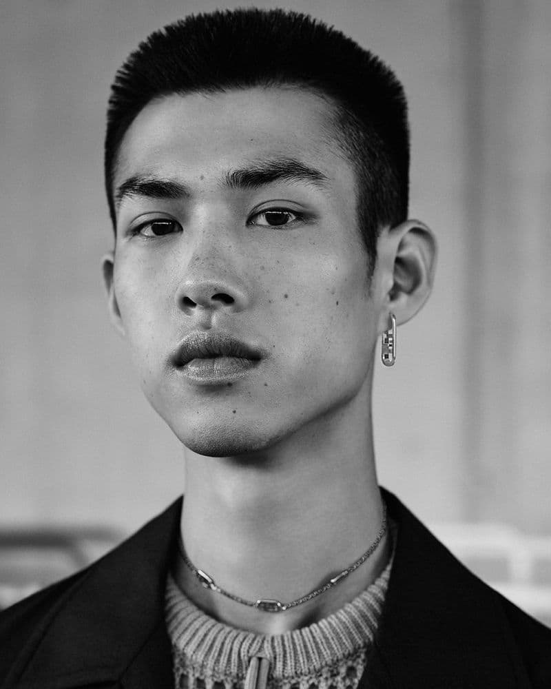 Close-up black and white portrait of a young man wearing a patterned knit top and dark jacket, featuring a silver chain necklace.