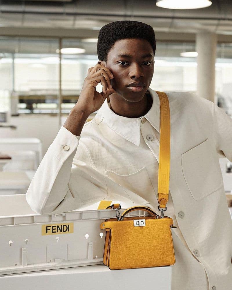 Portrait of a young adult male posing with a yellow Fendi handbag in an indoor setting with neutral light and wearing a button-down shirt.