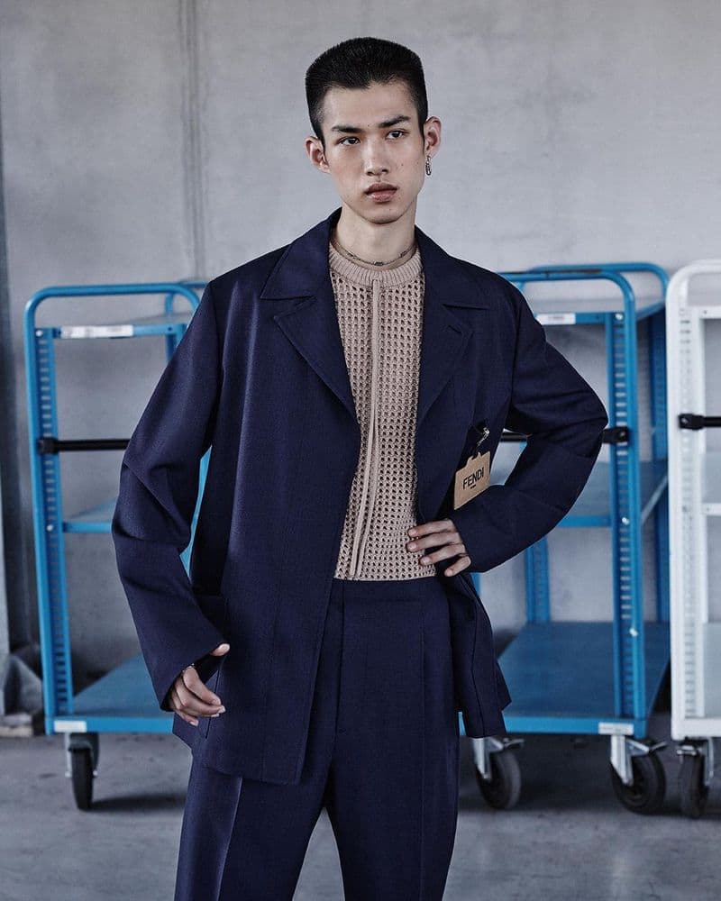 Fashion portrait of a young adult male model in a dark blue suit and beige top, standing in front of industrial utility carts.