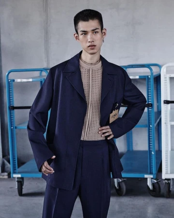 Fashion portrait of a young adult male model in a dark blue suit and beige top, standing in front of industrial utility carts.