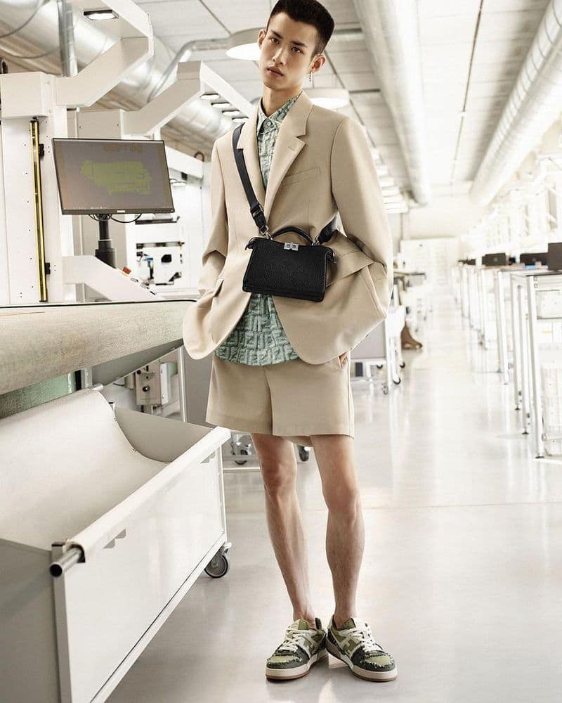 A young Asian man stands in a bright industrial setting wearing a beige suit, printed shirt, shorts, sneakers, and a black leather handbag.