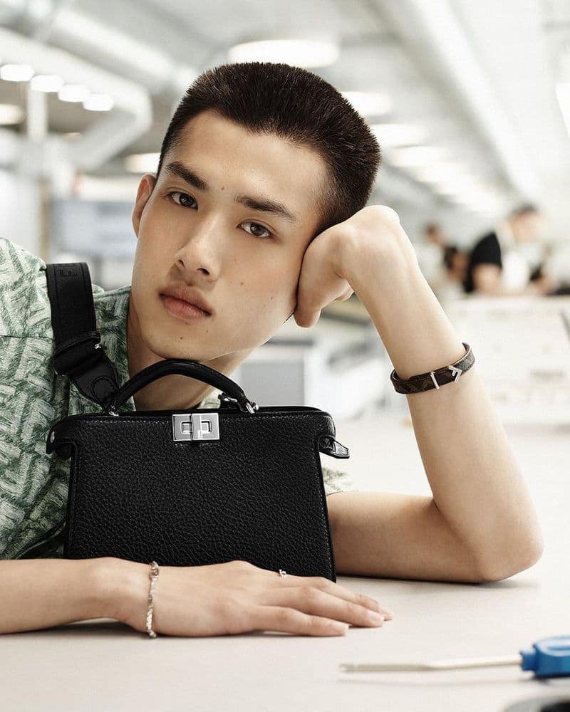 Stylish Asian male model poses indoors with a black leather handbag, showcasing fashion and style with a blurred background.