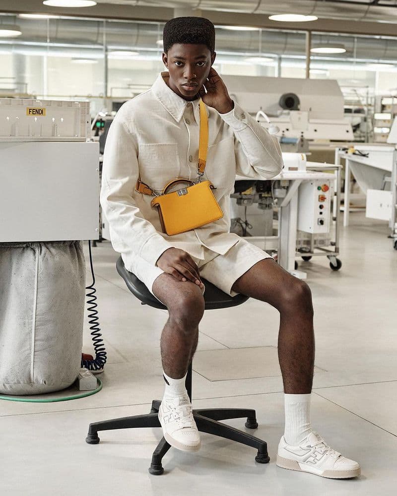 A young Black man poses in white linen shirt and shorts with a yellow purse, sitting on a stool in a bright industrial setting.