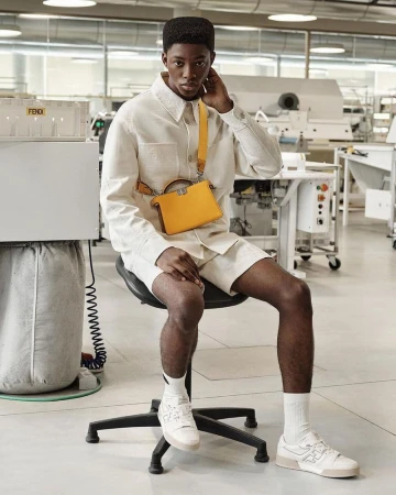 A young Black man poses in white linen shirt and shorts with a yellow purse, sitting on a stool in a bright industrial setting.