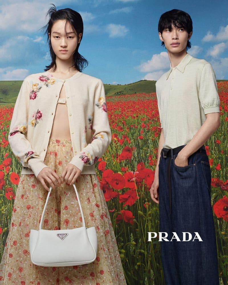 Two fashion models pose in a field of red poppies with a blue sky; the female model holds a white Prada bag. Prada logo is visible.