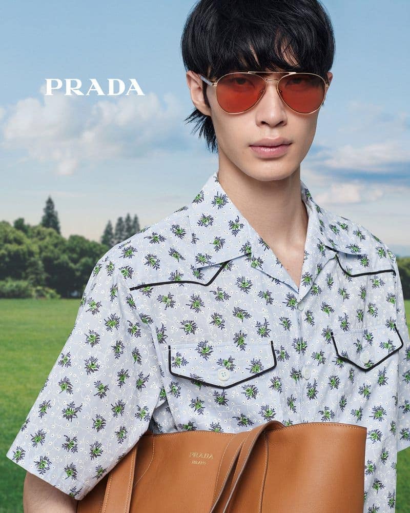 A fashionable young man wearing a patterned shirt and aviator sunglasses poses with a brown leather tote bag in a sunny outdoor setting.
