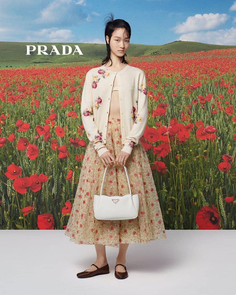 A young woman poses in a floral skirt and sweater in a field of red flowers, holding a white Prada handbag under a blue sky.