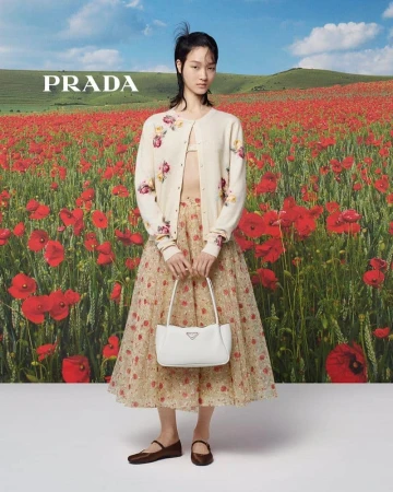 A young woman poses in a floral skirt and sweater in a field of red flowers, holding a white Prada handbag under a blue sky.