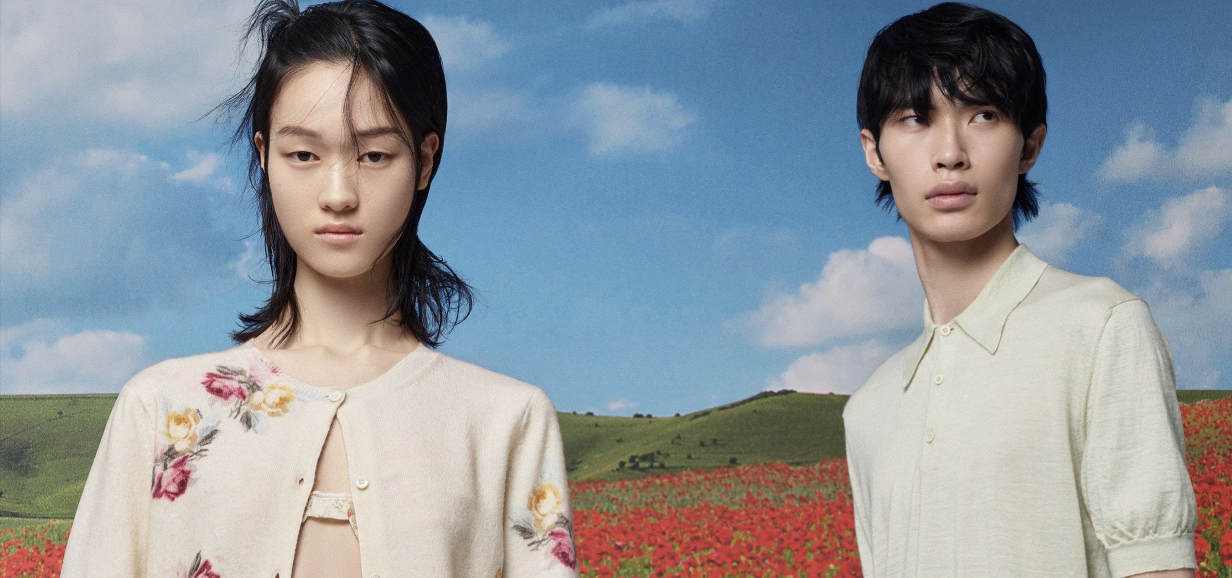 Two Asian models pose outdoors in front of a vibrant field of red flowers and rolling green hills under a bright blue sky with scattered clouds.