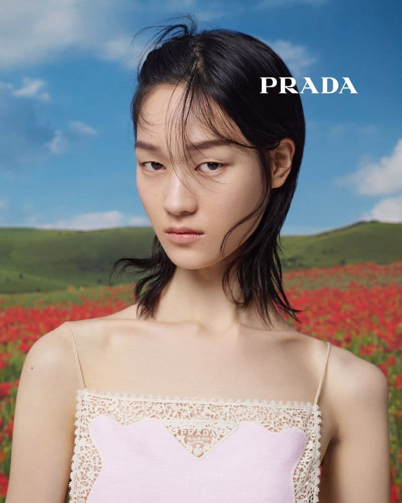 Asian model with dark hair, pale pink top, and lace trim poses in a field of red flowers under a blue sky for a fashion advertisement.