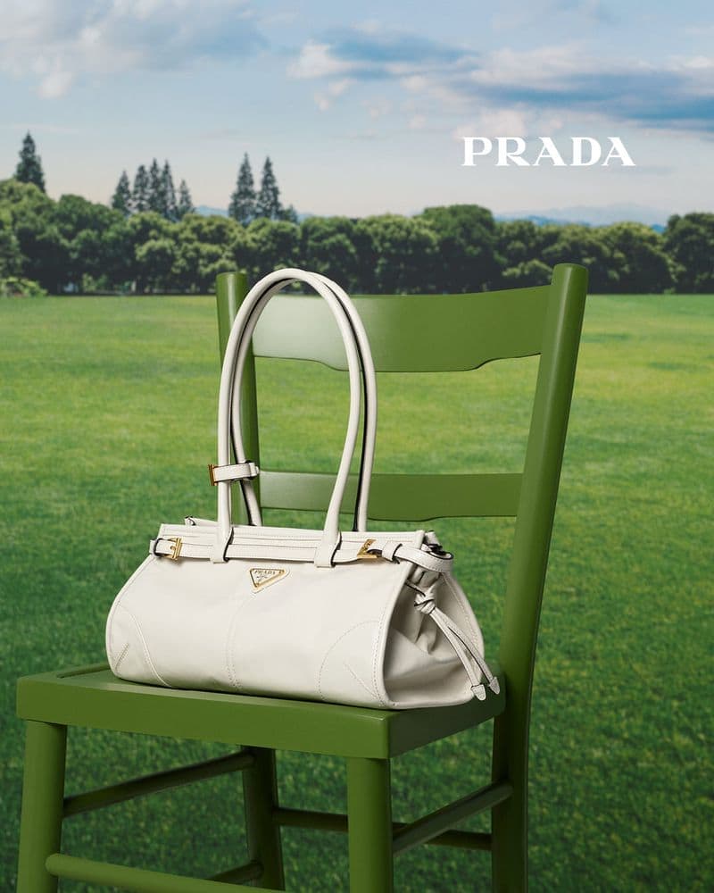 White leather Prada handbag with gold hardware sitting on a green wooden chair, set in a lush green field under a blue sky.