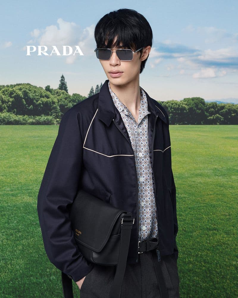 A stylish young man with sunglasses, wearing a jacket and carrying a black messenger bag, poses in front of a green landscape.