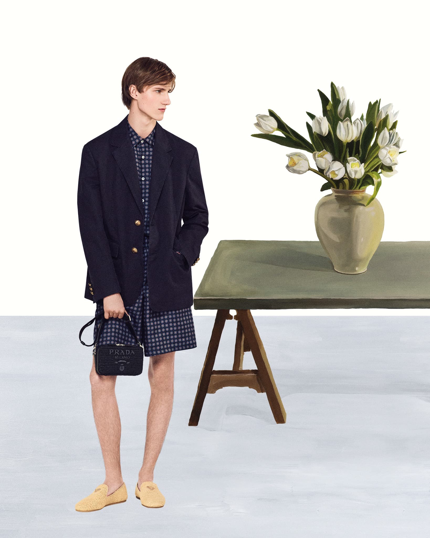 Male fashion model poses in navy blazer, print shirt and shorts, holding black Prada purse near tulips and table.