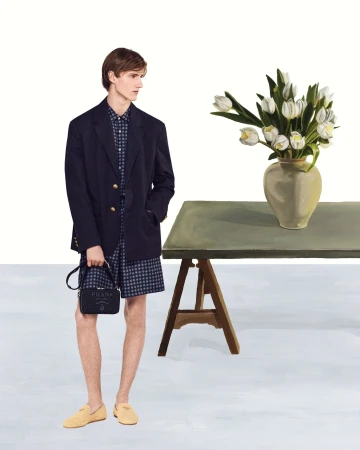 Male fashion model poses in navy blazer, print shirt and shorts, holding black Prada purse near tulips and table.