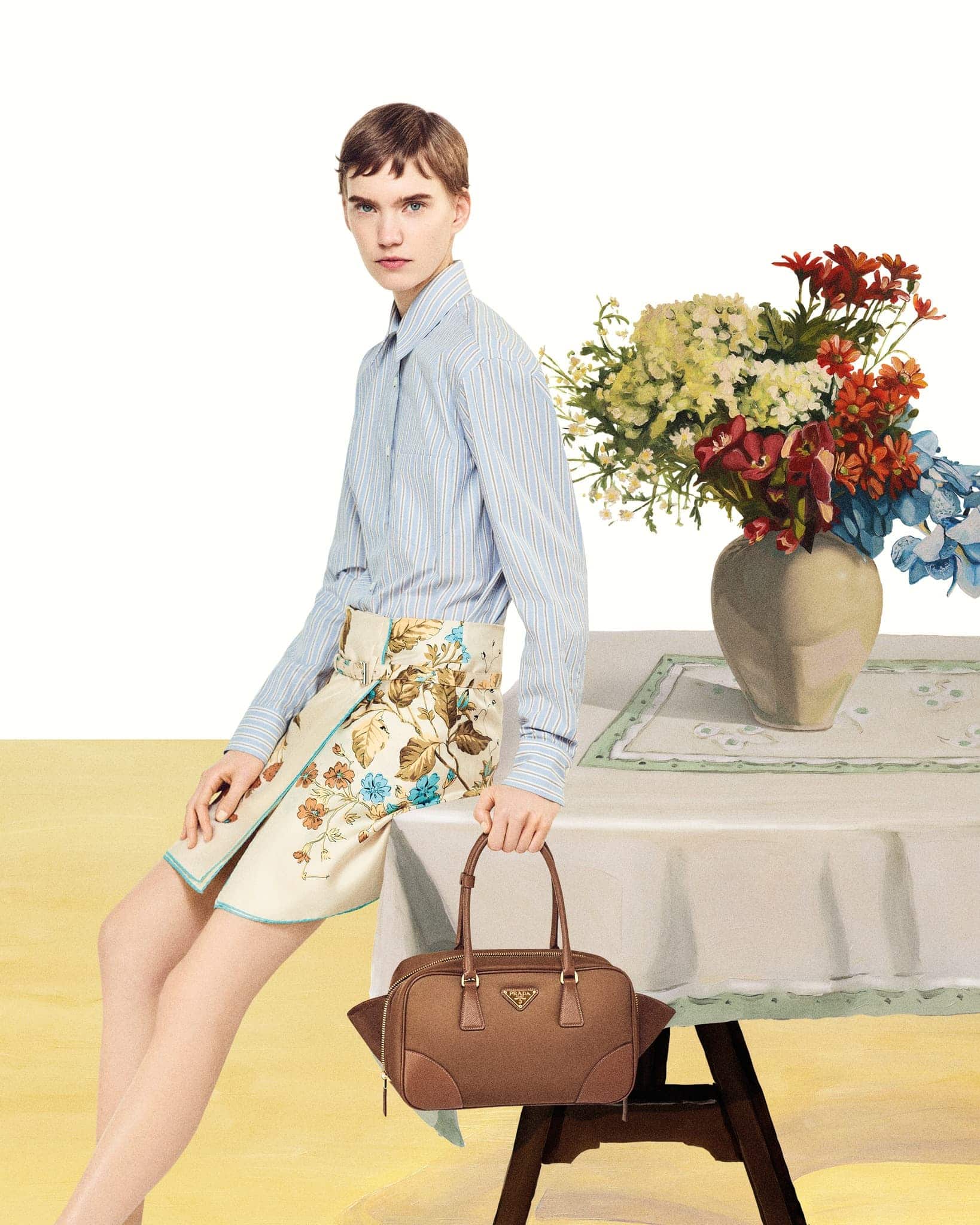Adult female seated next to table with flowers, wearing a skirt and blouse, holding a brown leather purse with Prada logo.