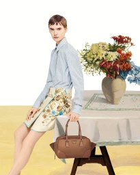 Model sitting on a table next to a floral vase, holding a brown leather handbag, for Prada Summer 2024 campaign