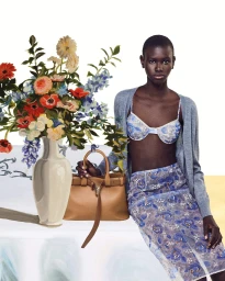 Model wearing a blue floral bra and matching skirt with a grey cardigan holding a tan leather bag next to a flower vase on a table