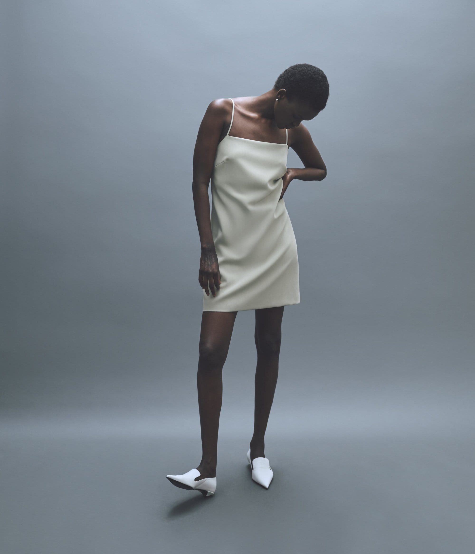 A slender Black woman poses in a studio, showcasing a pale-yellow sleeveless mini dress and white pointed-toe shoes against a gray backdrop.