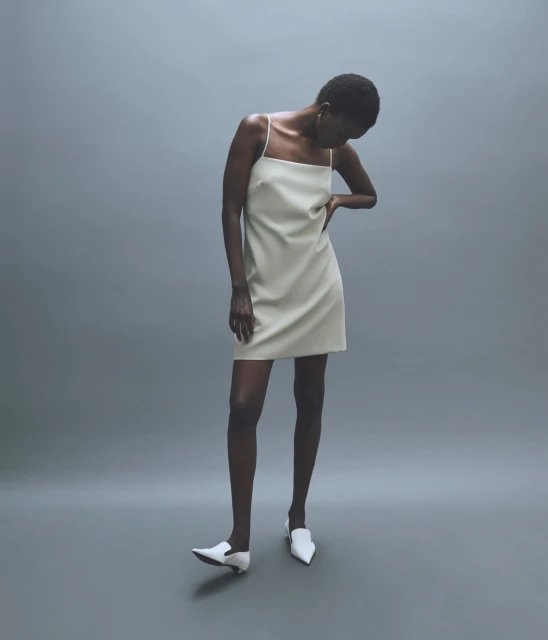 A slender Black woman poses in a studio, showcasing a pale-yellow sleeveless mini dress and white pointed-toe shoes against a gray backdrop.