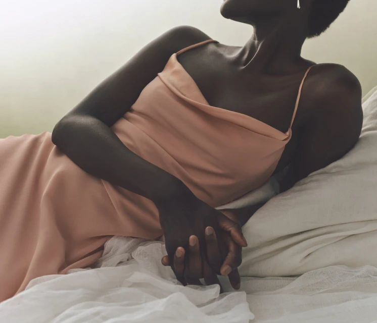 A dark-skinned woman in a peach-colored dress lying on a white bed. Soft, diffused light highlights the textures of skin and fabric.