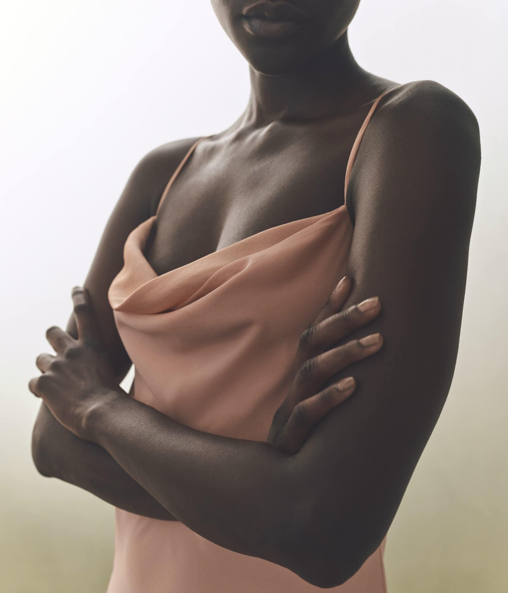 Close-up studio shot of a dark-skinned adult female in a peach-colored sleeveless dress, posing with her arms crossed against a hazy white backdrop.