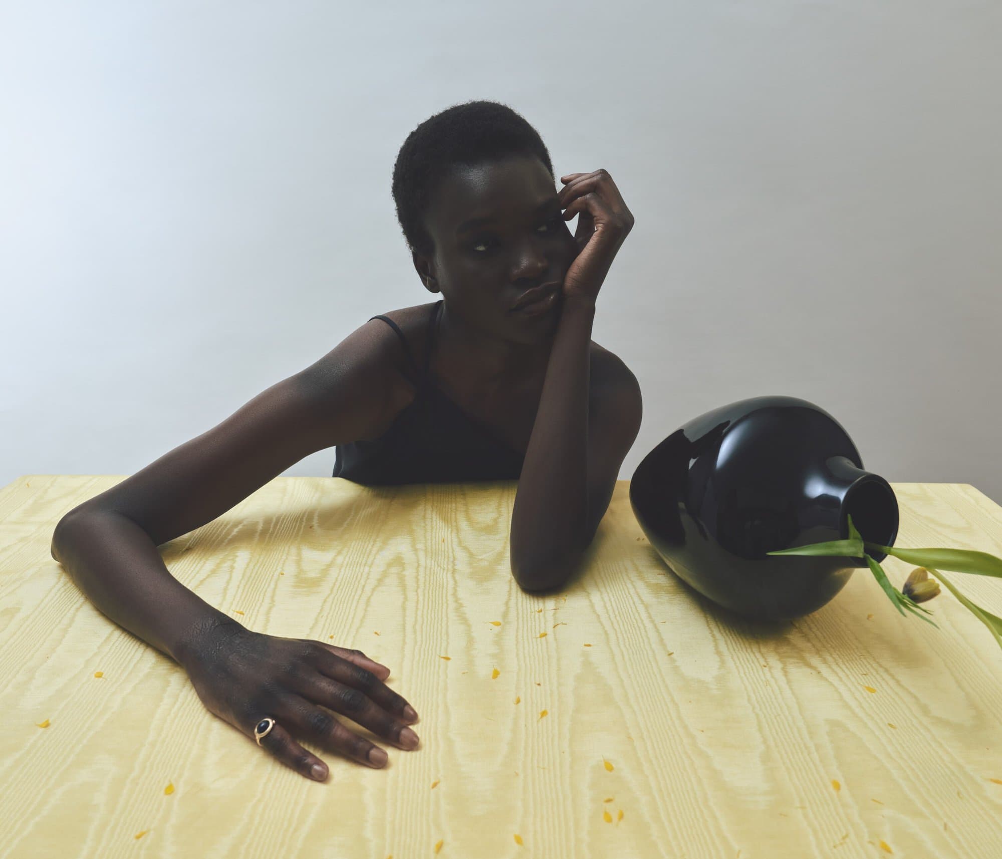 Portrait of a dark-skinned woman with short hair resting her head on her hand at a wooden table with a black vase and greenery.