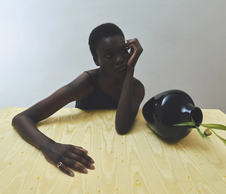Portrait of a dark-skinned woman with short hair resting her head on her hand at a wooden table with a black vase and greenery.