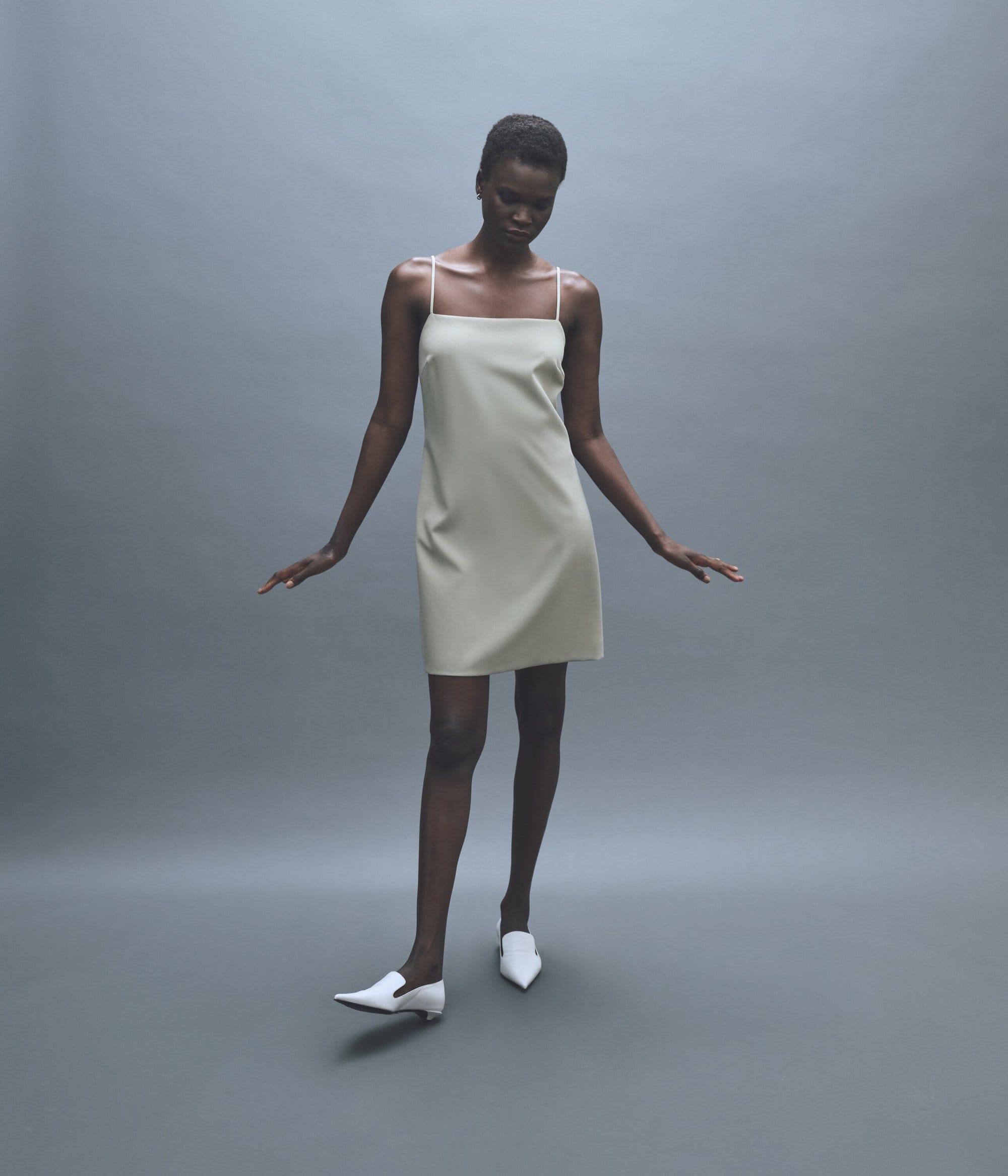 Dark-skinned fashion model showcases a cream-colored mini dress with spaghetti straps and white pointed-toe heeled shoes in a studio setting.