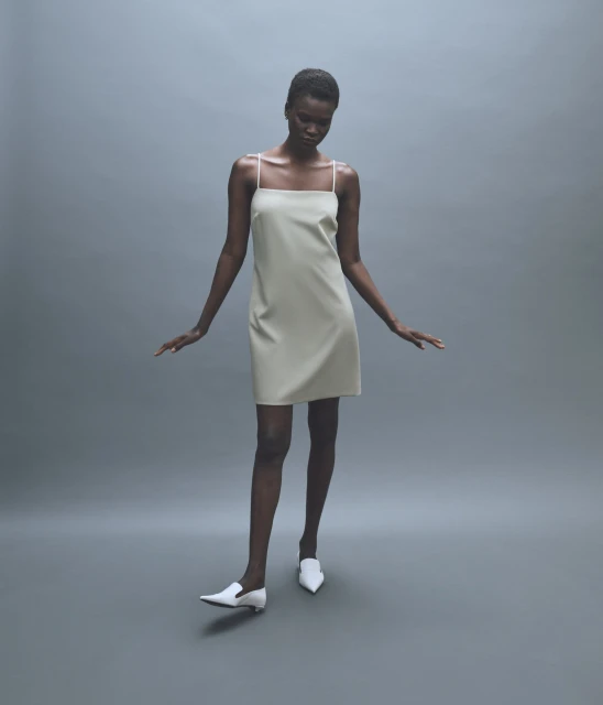 Dark-skinned fashion model showcases a cream-colored mini dress with spaghetti straps and white pointed-toe heeled shoes in a studio setting.