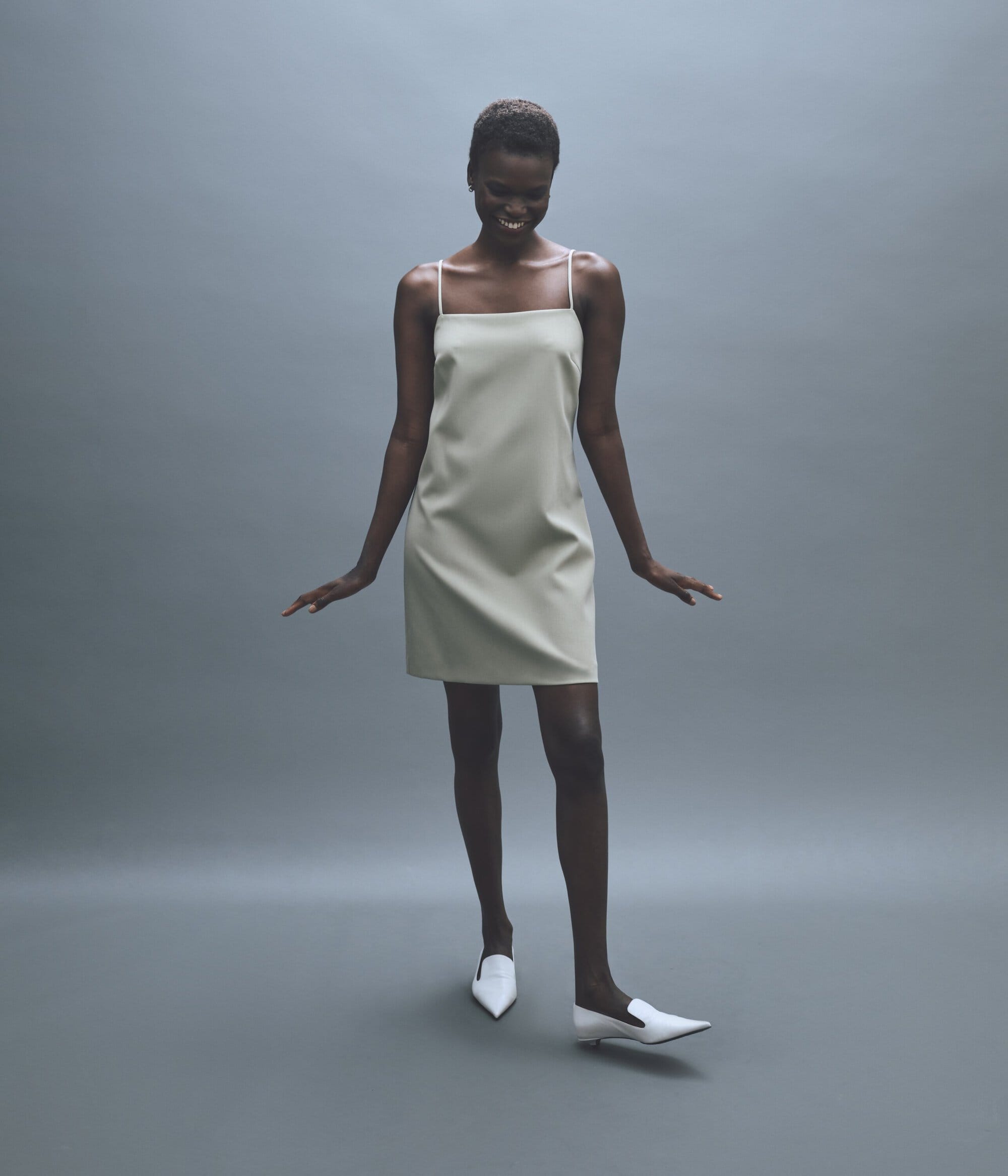 Model wearing a light slip dress and white pointed mules posing against a grey studio backdrop