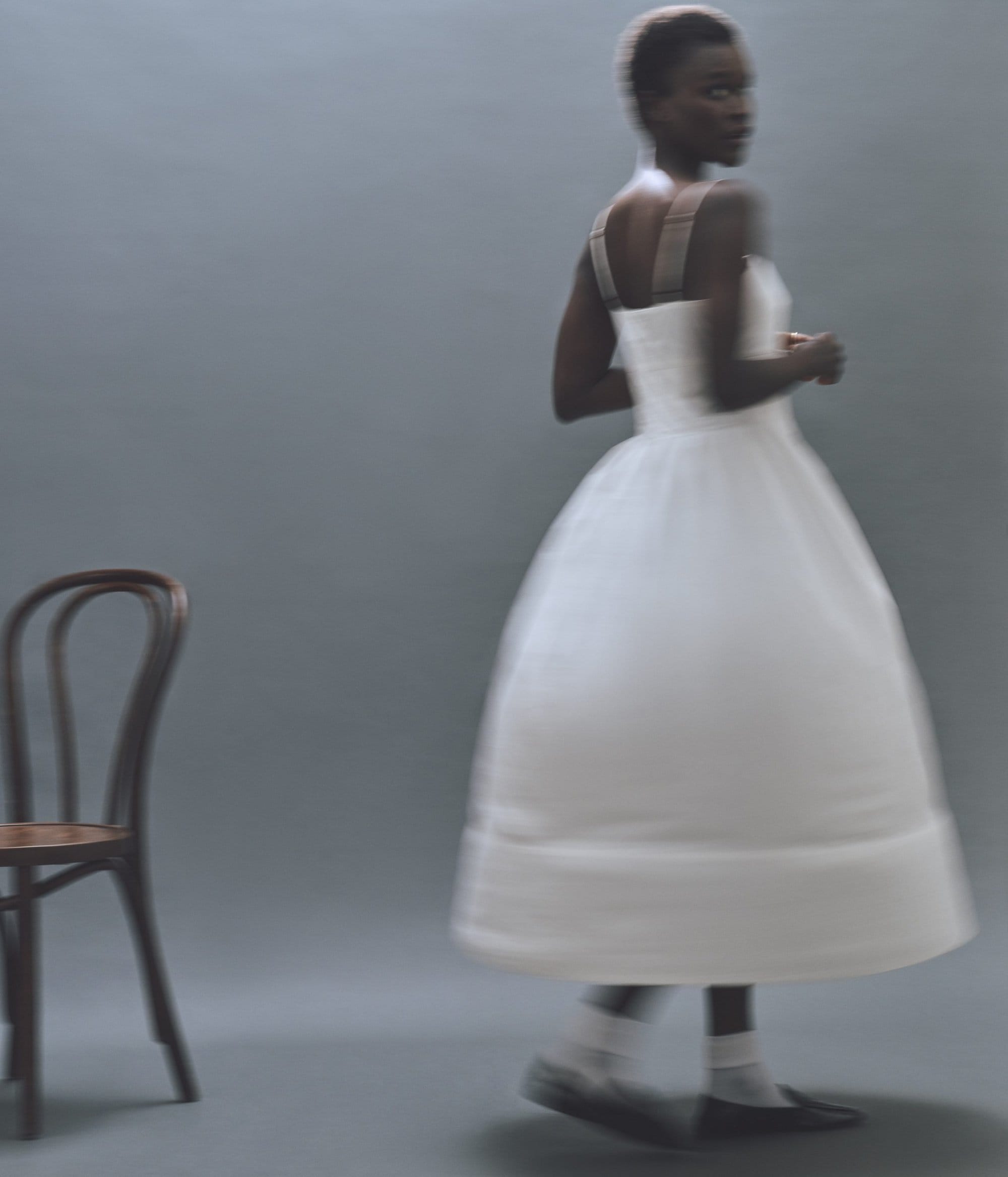 Fashion shot of an African woman in a white dress looking over her shoulder, set against a gray background with a wooden chair.