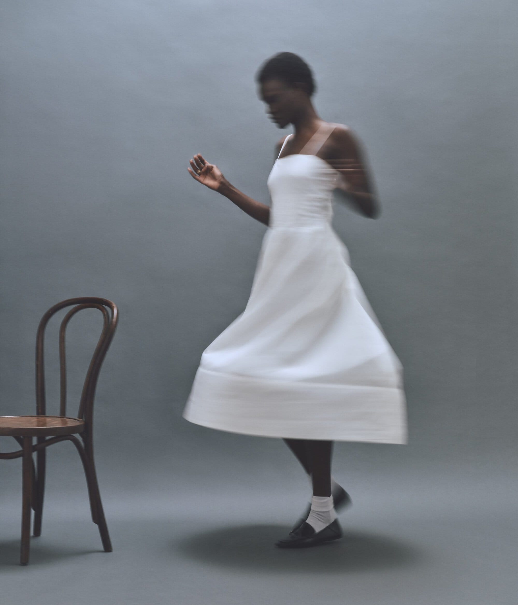 A blurred motion portrait of an African-American woman in a white dress with a chair in a grey indoor studio setting.