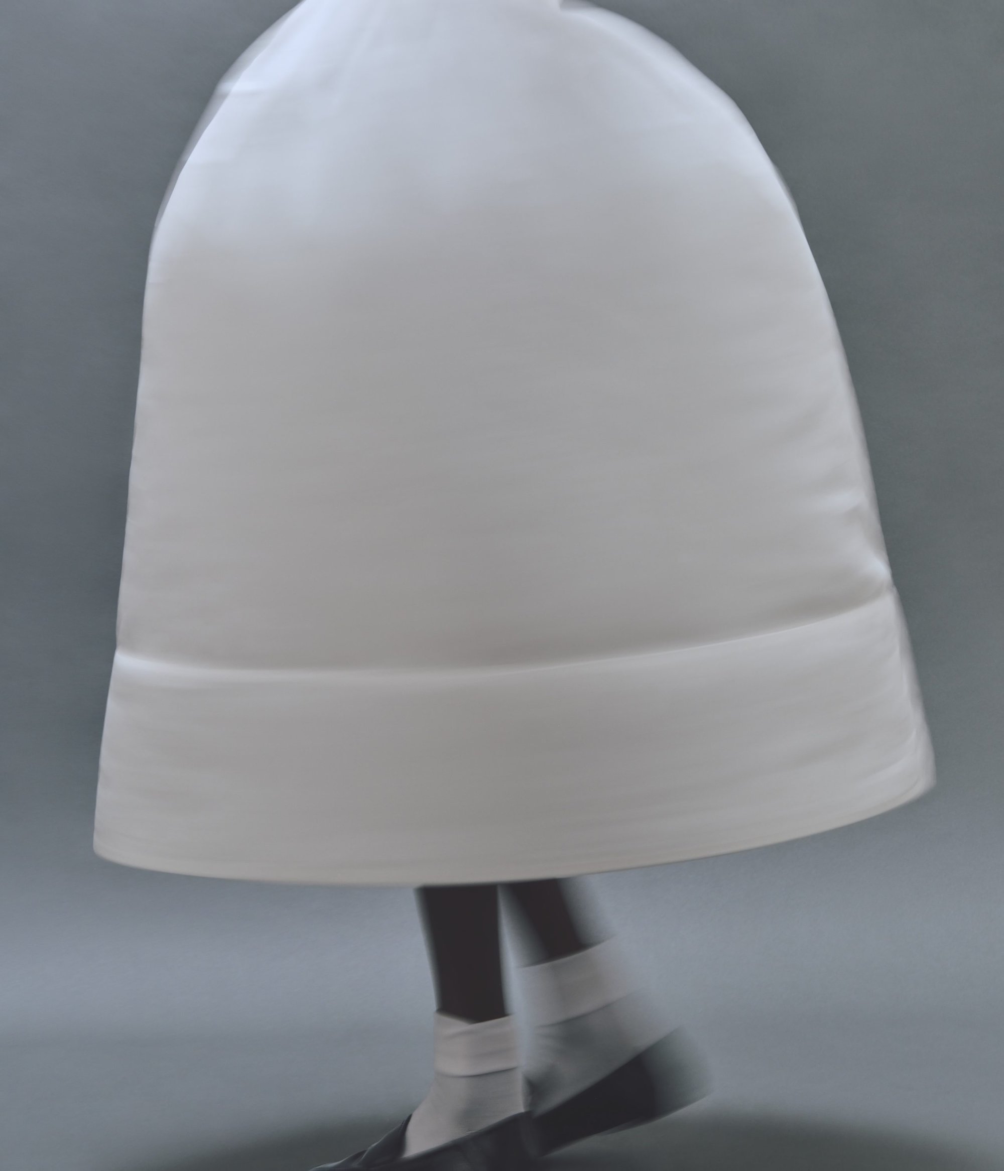 Model wearing a structured white bell-shaped skirt with white socks and dark shoes against a grey background