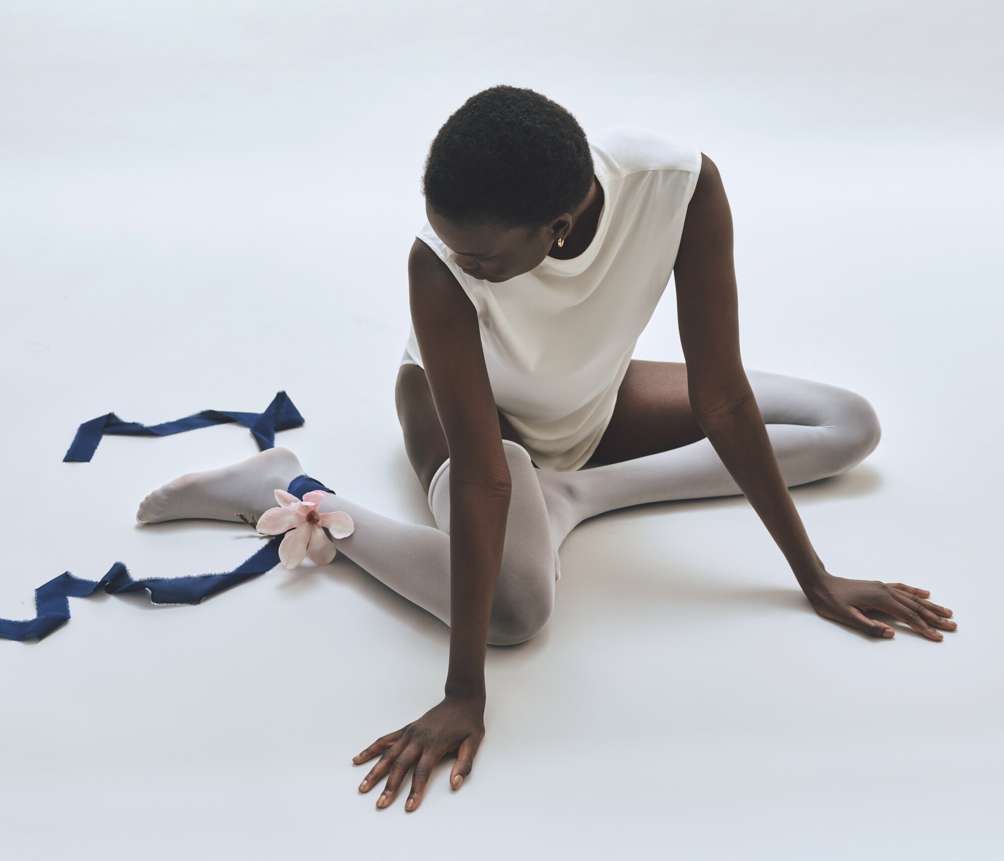 Elegant studio shot of a Black woman in white top and tights, seated with a ribbon and flower accenting her leg, against a white backdrop.