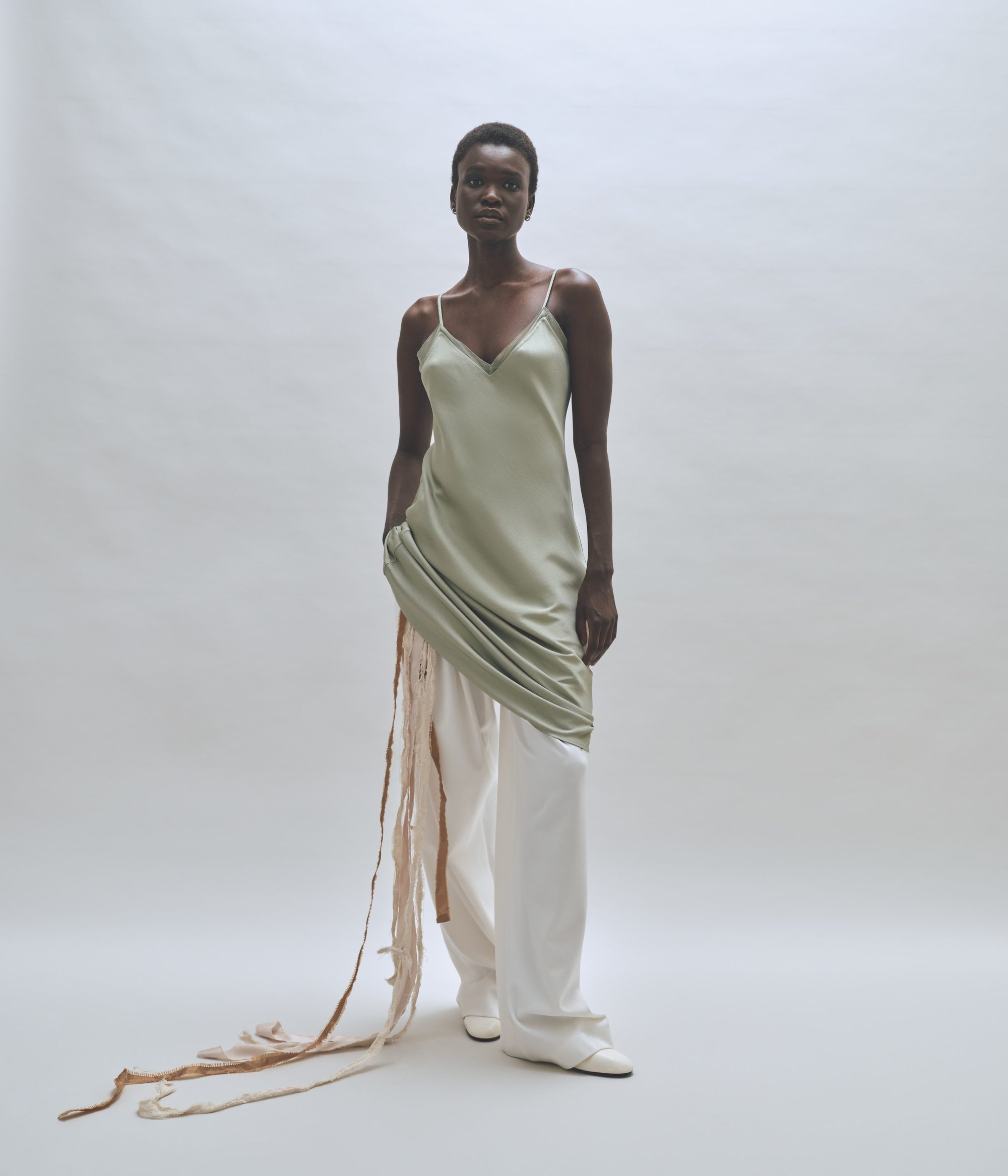 Full-length studio shot of an African-American female fashion model wearing a slip dress over white pants and matching pointed-toe shoes.