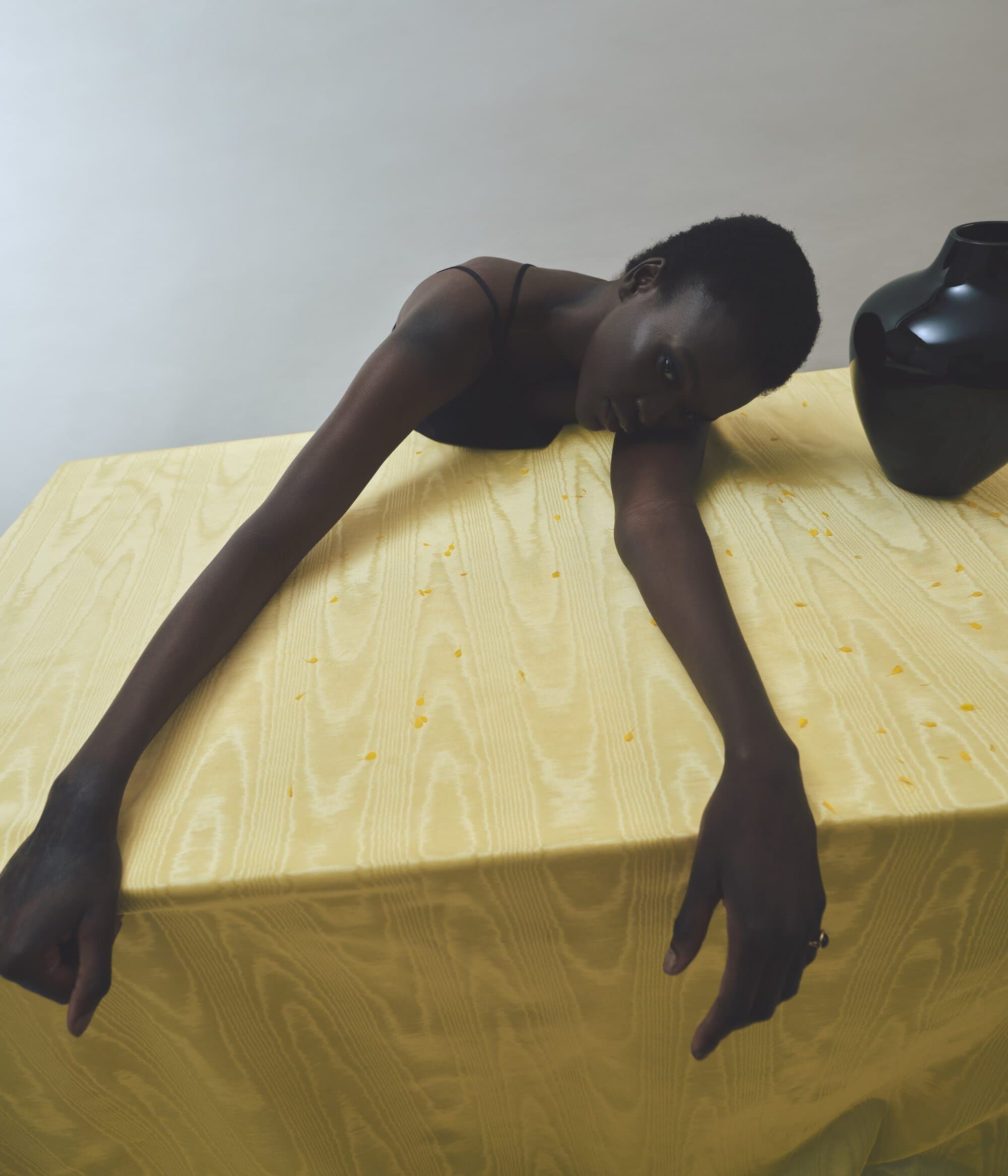 Fashionable studio portrait of a dark-skinned woman leaning across a yellow cloth-covered table next to a dark vase against a plain wall.