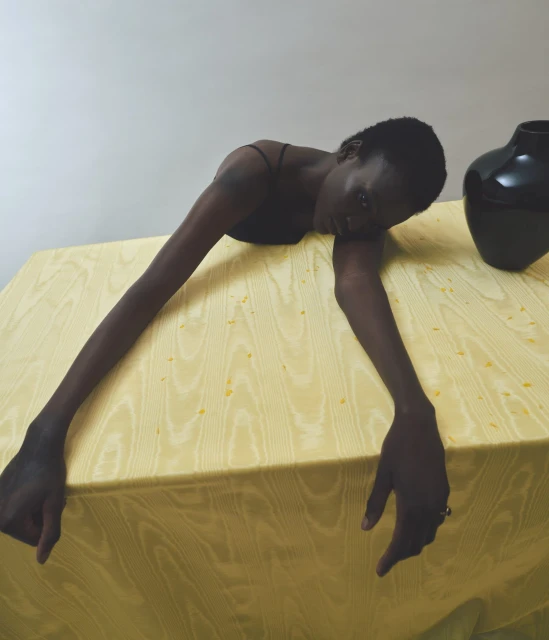 Fashionable studio portrait of a dark-skinned woman leaning across a yellow cloth-covered table next to a dark vase against a plain wall.