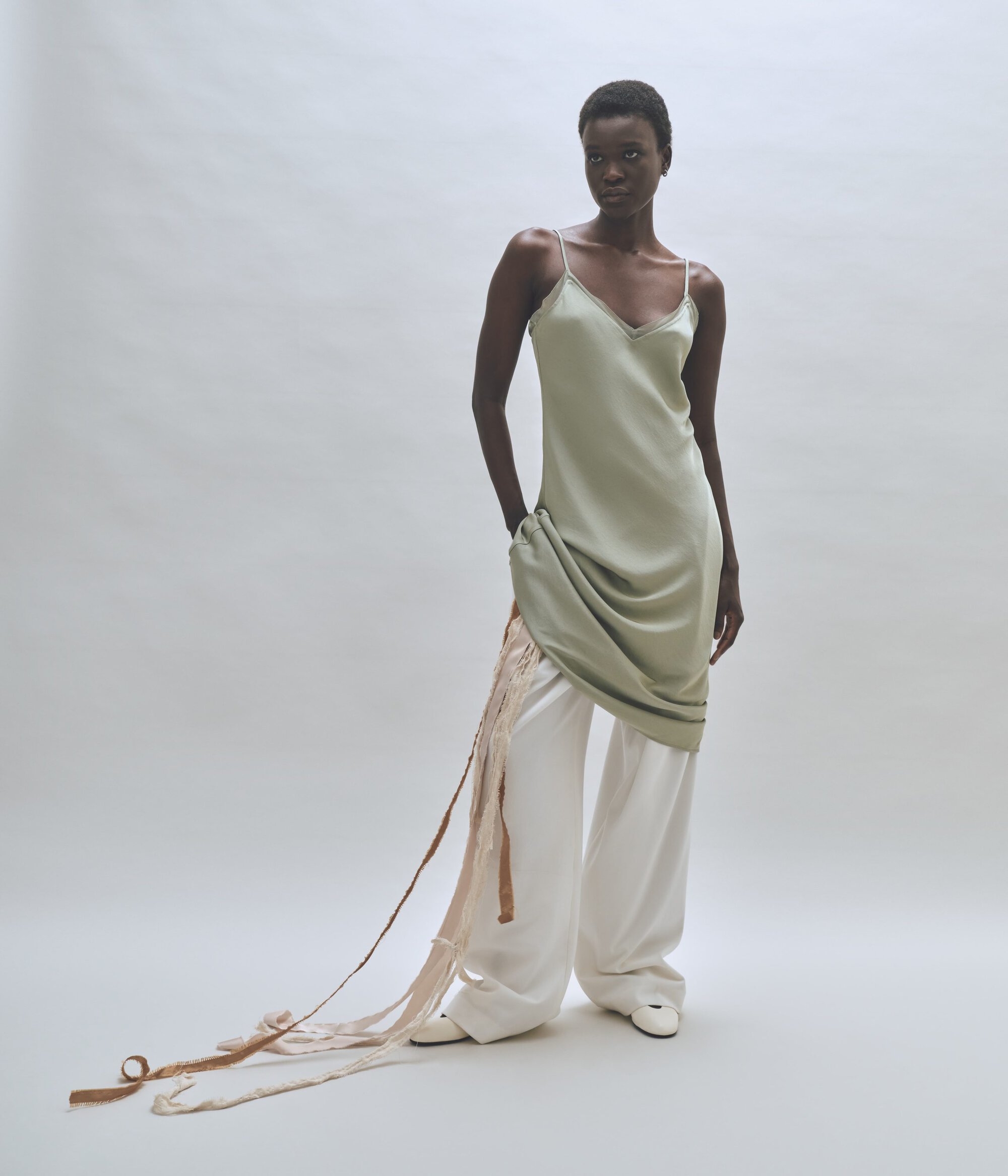 Model wearing a sage green slip dress and white trousers against a neutral background for Rue Sophie.