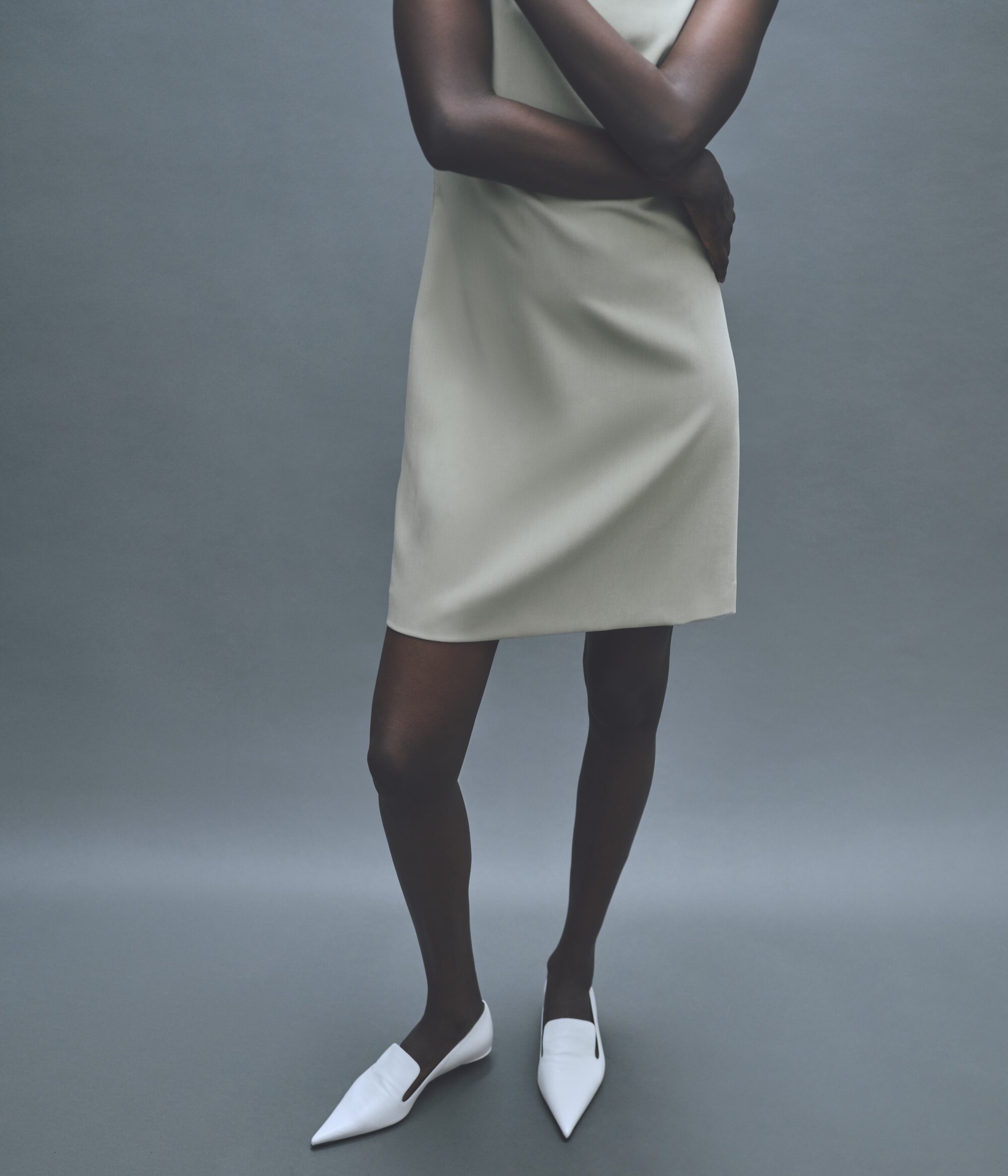 A slender, dark-skinned woman in a cream-colored dress and white flats stands against a neutral gray backdrop in a studio fashion shot.