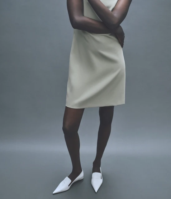 A slender, dark-skinned woman in a cream-colored dress and white flats stands against a neutral gray backdrop in a studio fashion shot.