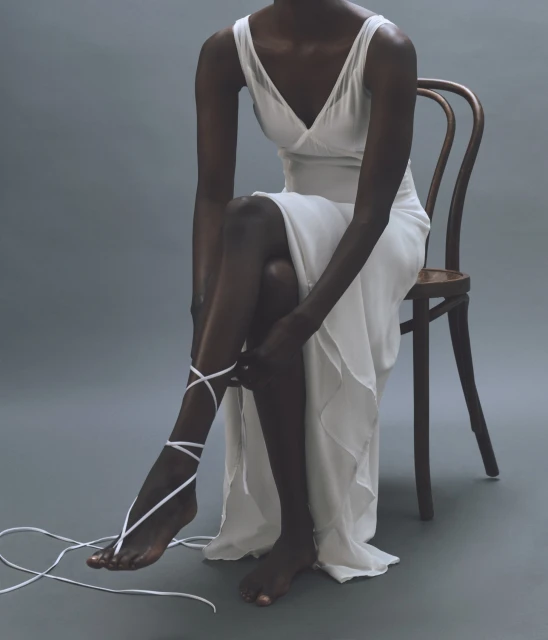 Dark-skinned woman sitting on a chair, tying white fabric around her leg, wearing a white dress, against a grey background.