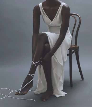 Dark-skinned woman sitting on a chair, tying white fabric around her leg, wearing a white dress, against a grey background.