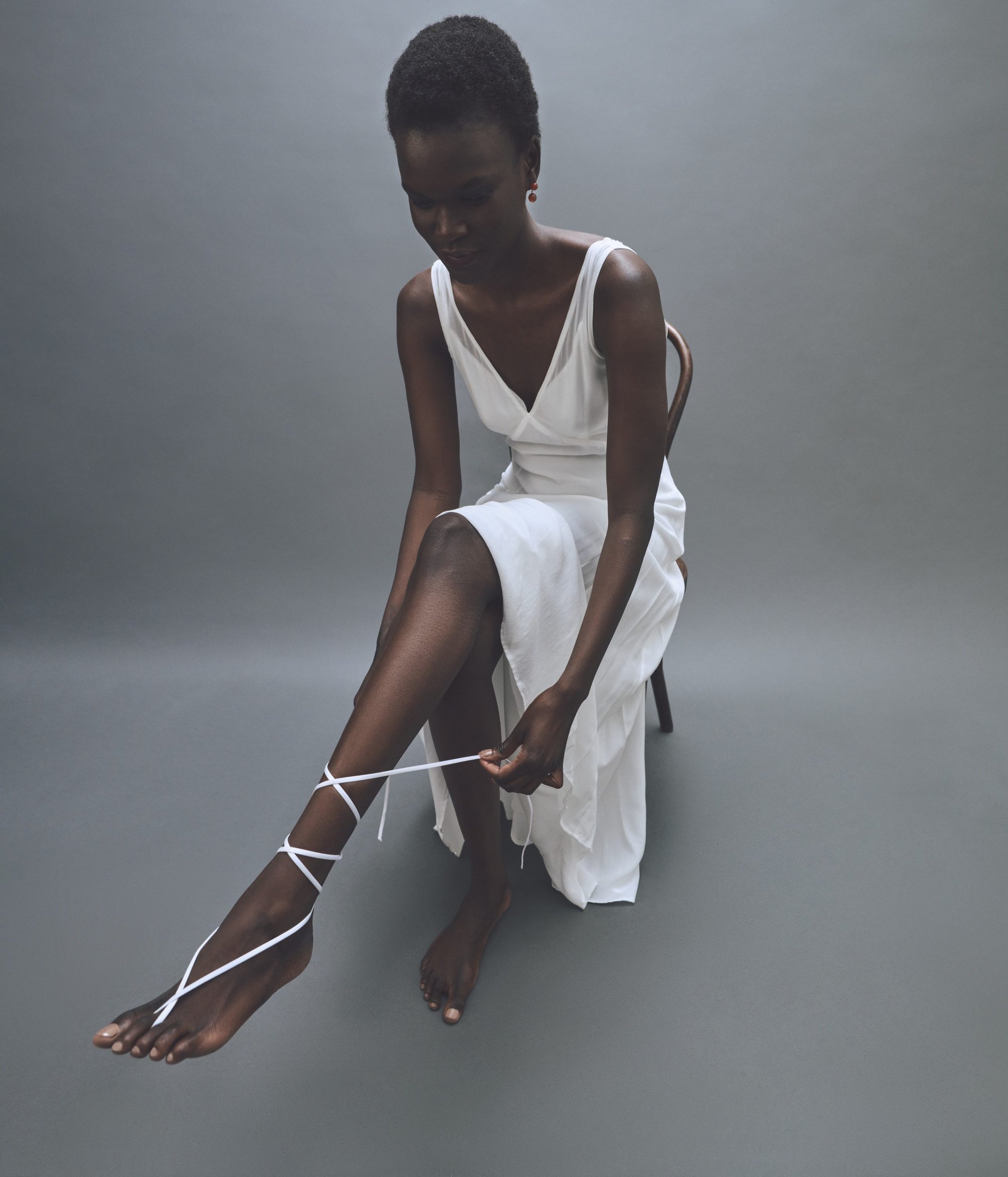 Elegant Black woman in a white dress sits tying a white strap around her ankle in a minimalist photo studio setting with grey backdrop.