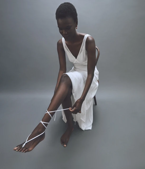Elegant Black woman in a white dress sits tying a white strap around her ankle in a minimalist photo studio setting with grey backdrop.