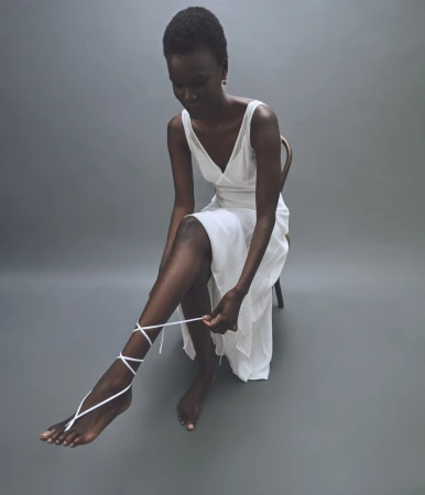 Elegant Black woman in a white dress sits tying a white strap around her ankle in a minimalist photo studio setting with grey backdrop.