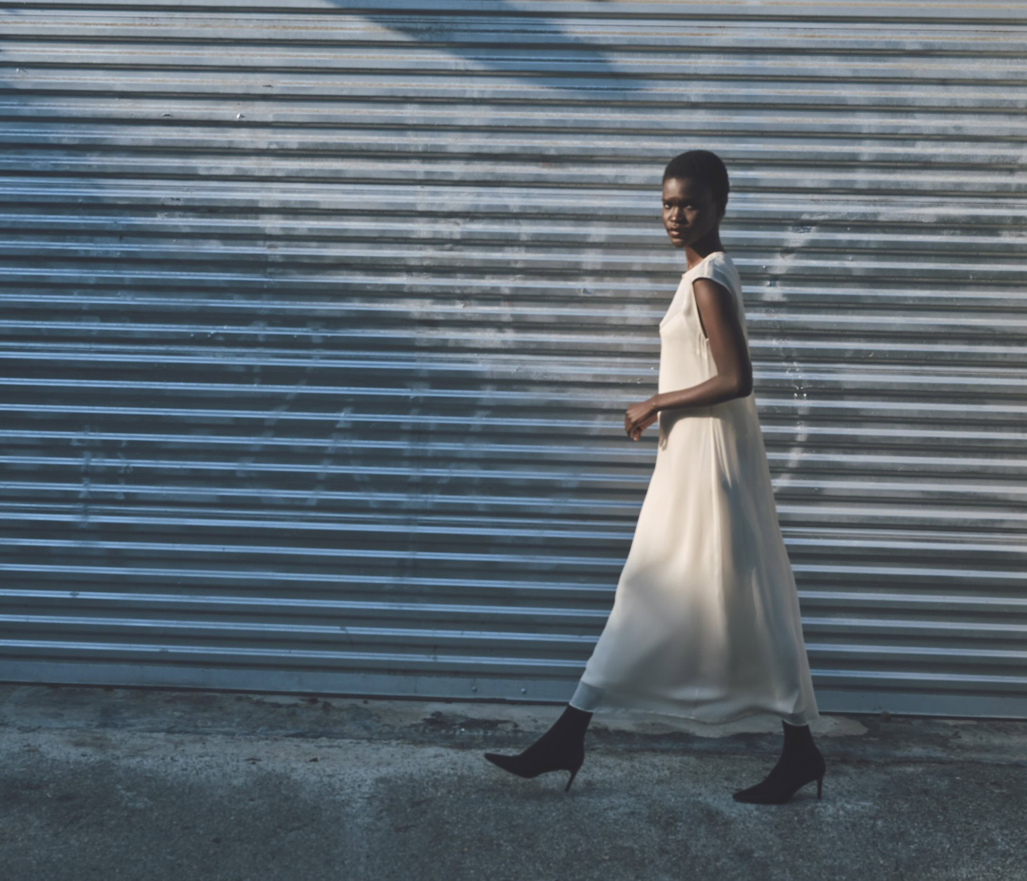 An African-American woman in a light-colored dress and black boots walks past a corrugated metal wall in an urban outdoor environment.