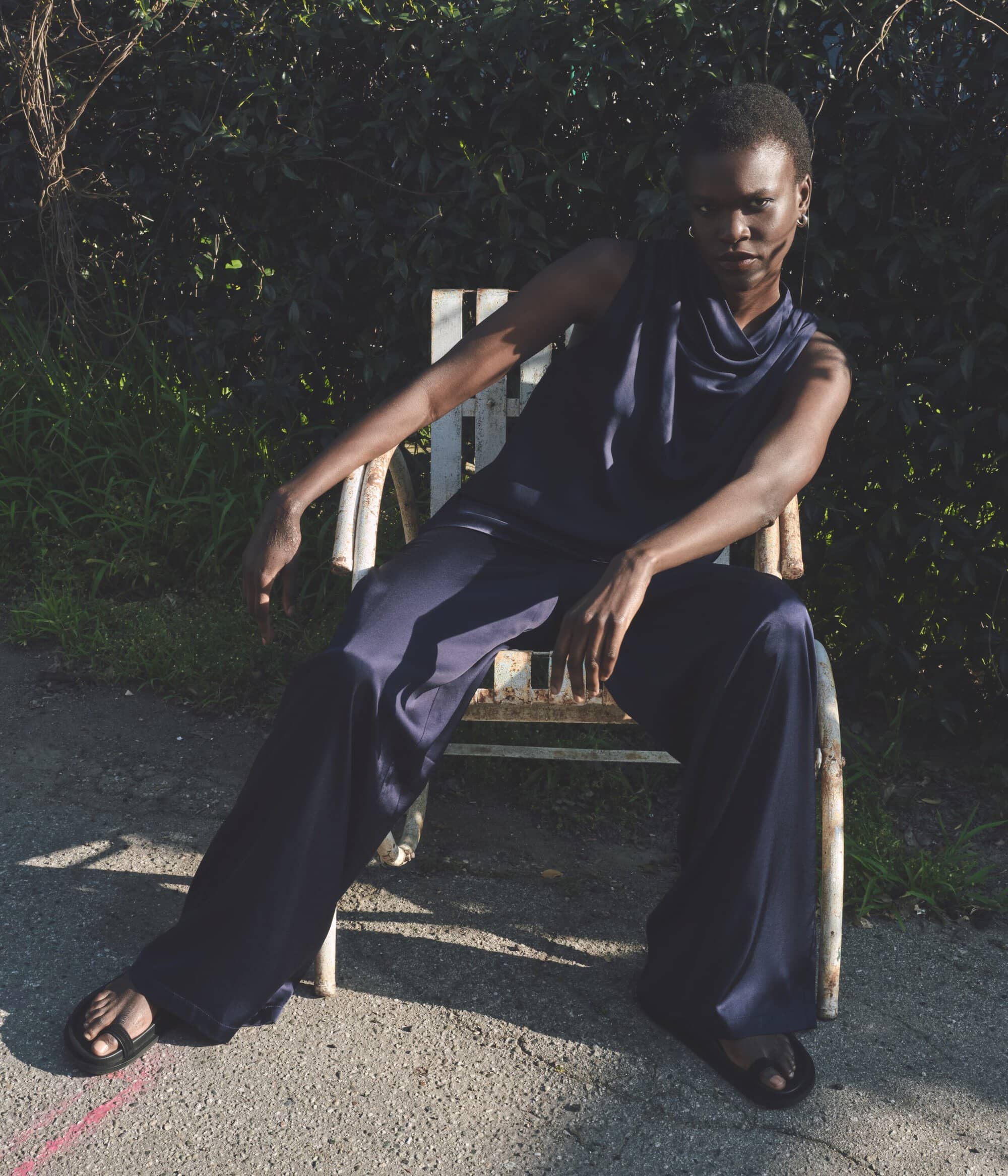 A fashionable Black woman in a navy jumpsuit sits on a white chair outdoors, posing in natural light with a serious facial expression.